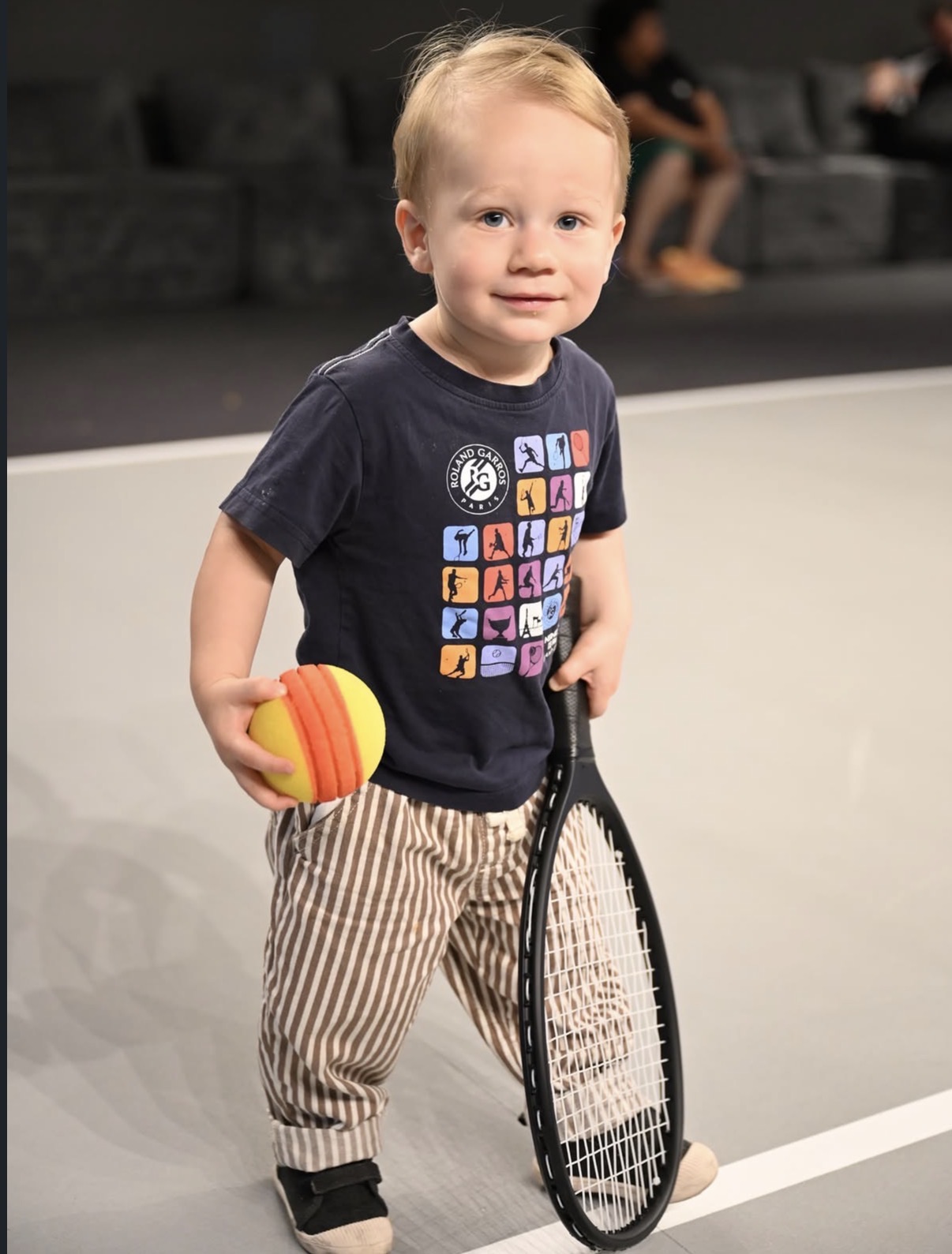 A young child holding a foam tennis ball and a small racket on the indoor court